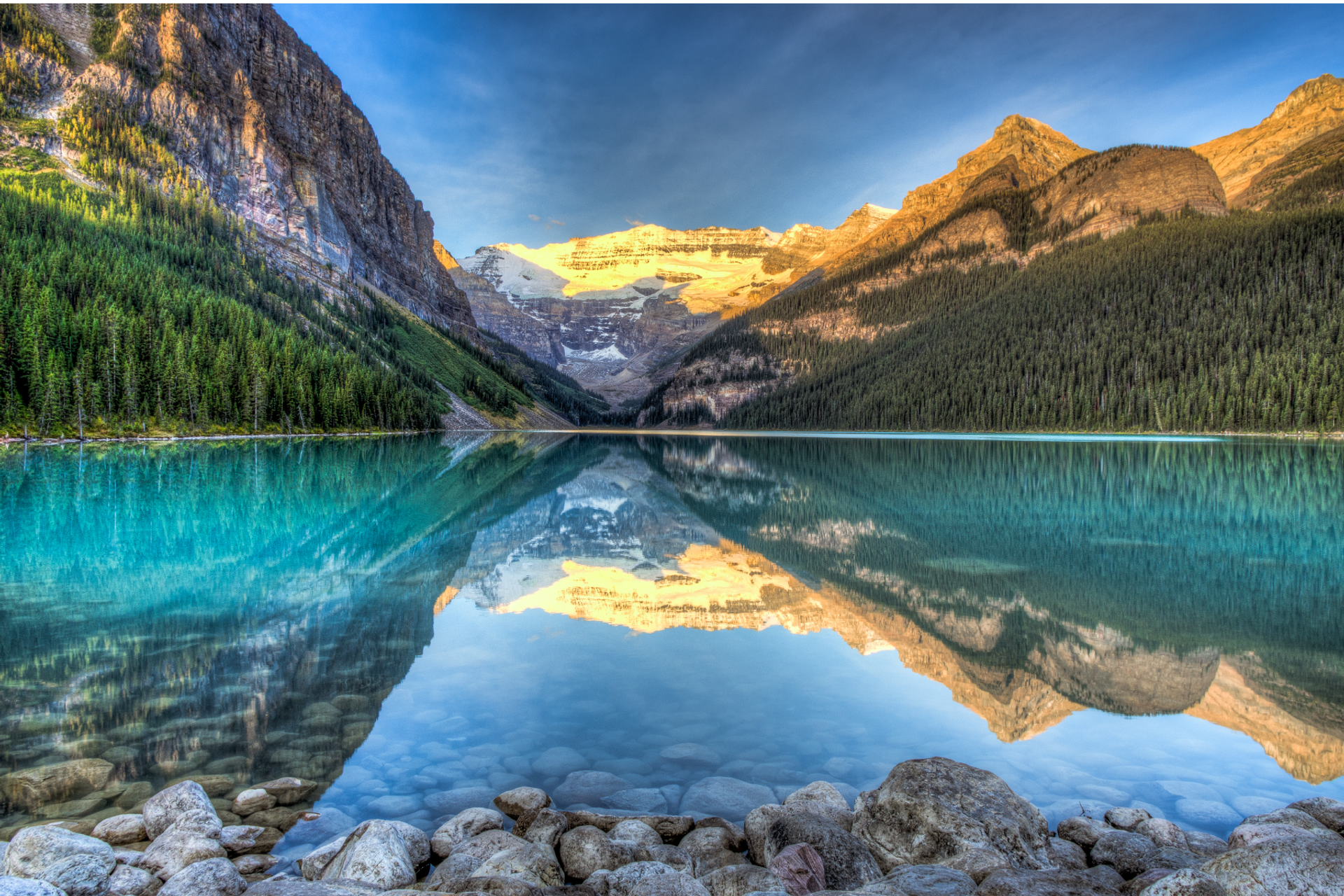 Crystal-clear lake reflecting mountains and forest under soft early morning light.