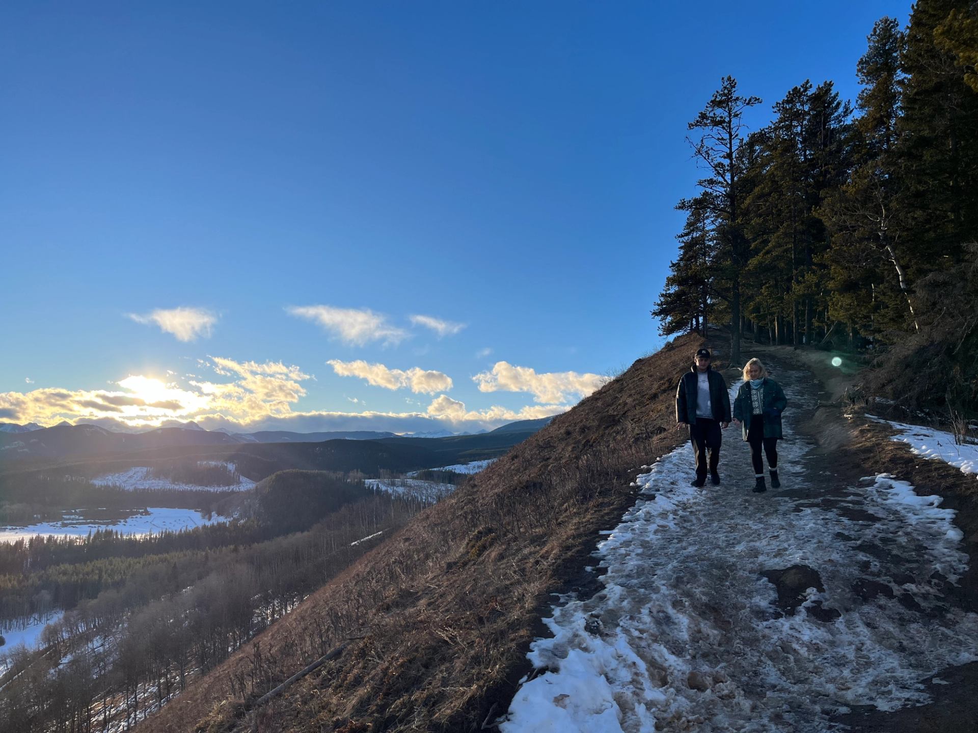 Two people hiking a snowy foothills trail with sunset lighting the valley.