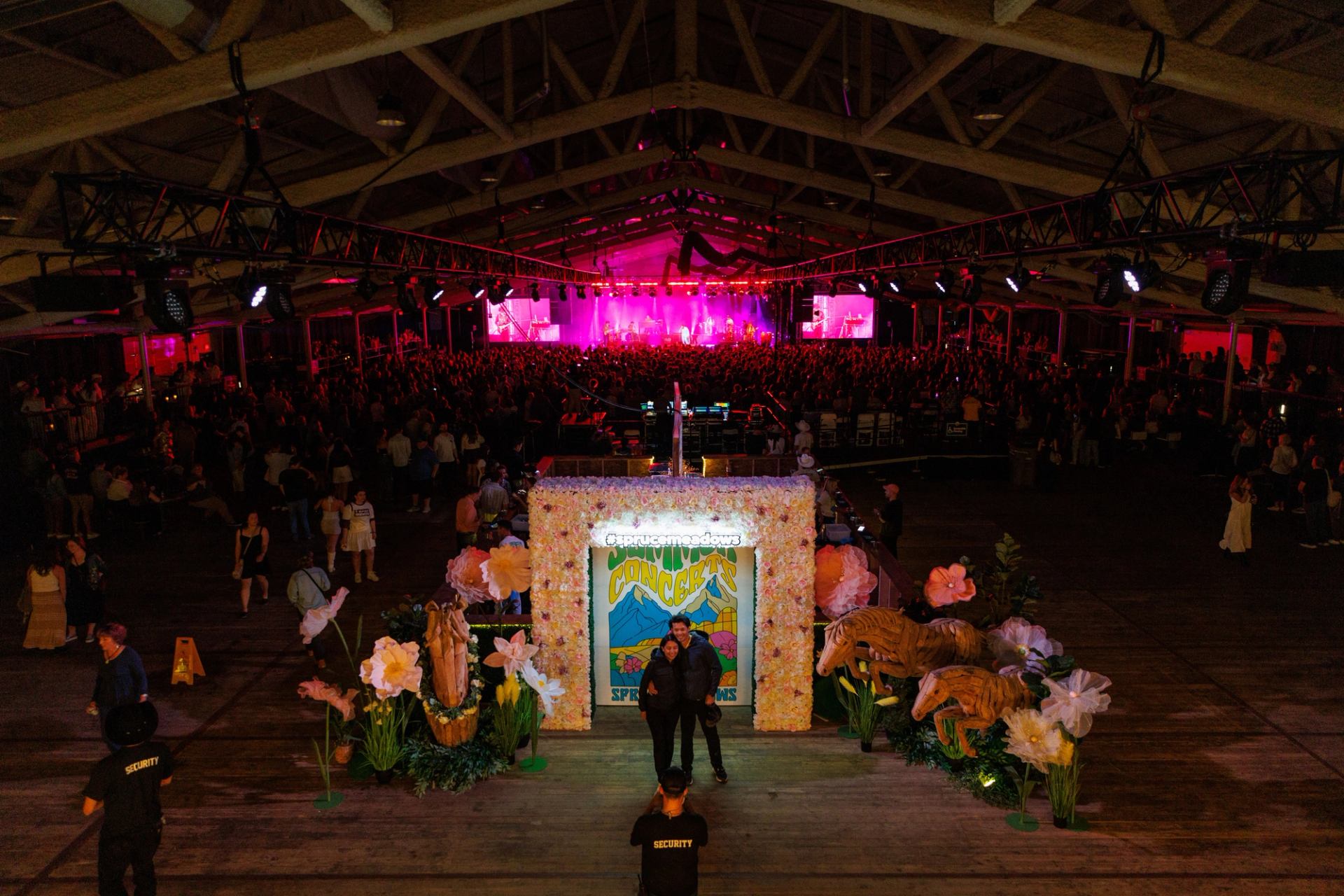 Indoor concert crowd faces lit stage, with floral photo arch and security guard foreground.