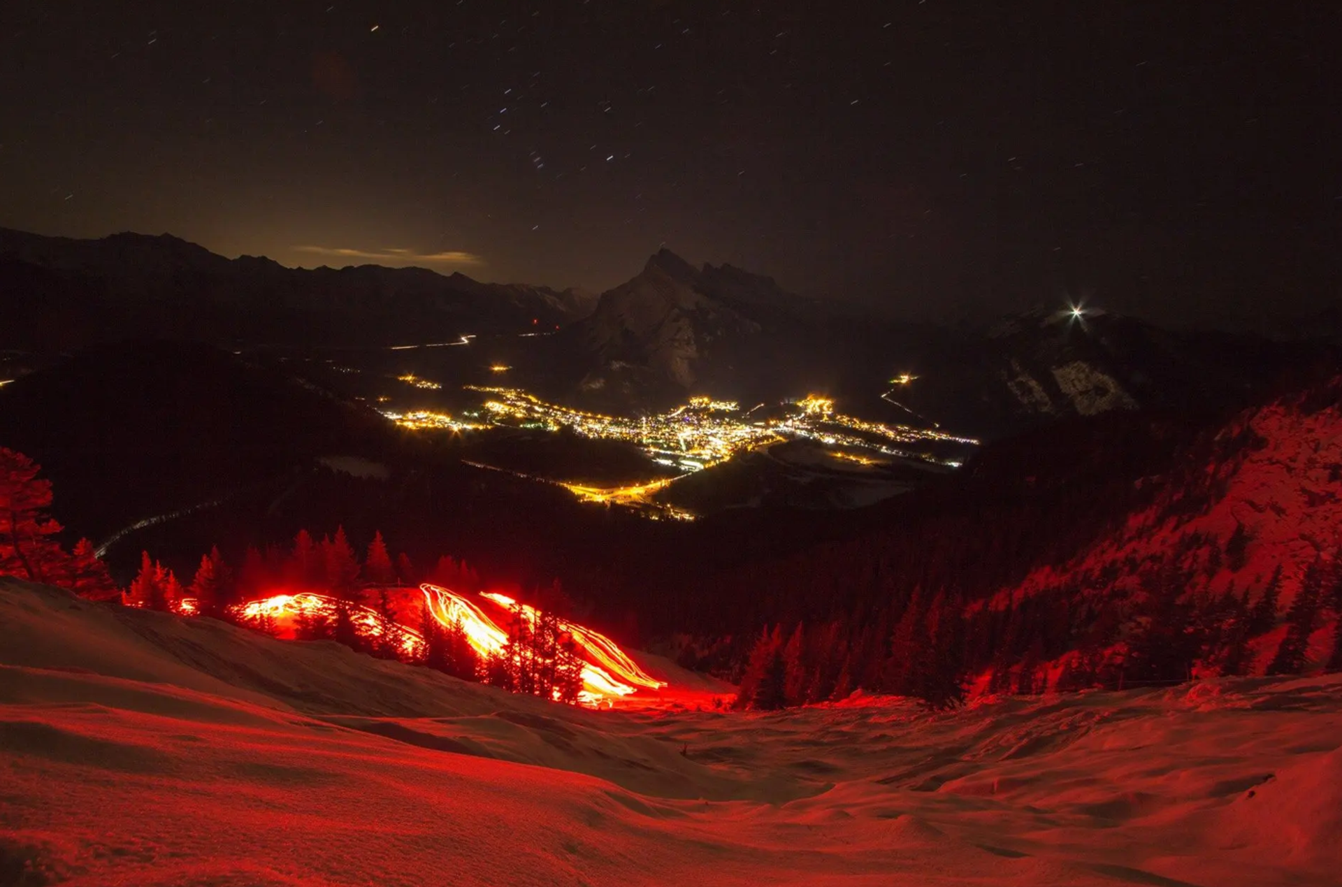 Glowing red torchlight trails on snowy slopes with a mountain town illuminated at night.