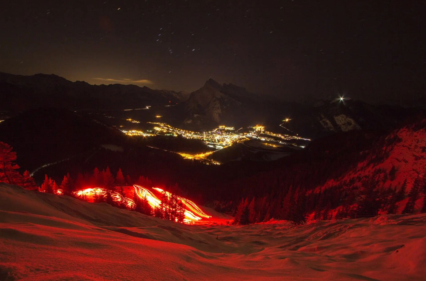 Glowing red torchlight trails on snowy slopes with a mountain town illuminated at night.