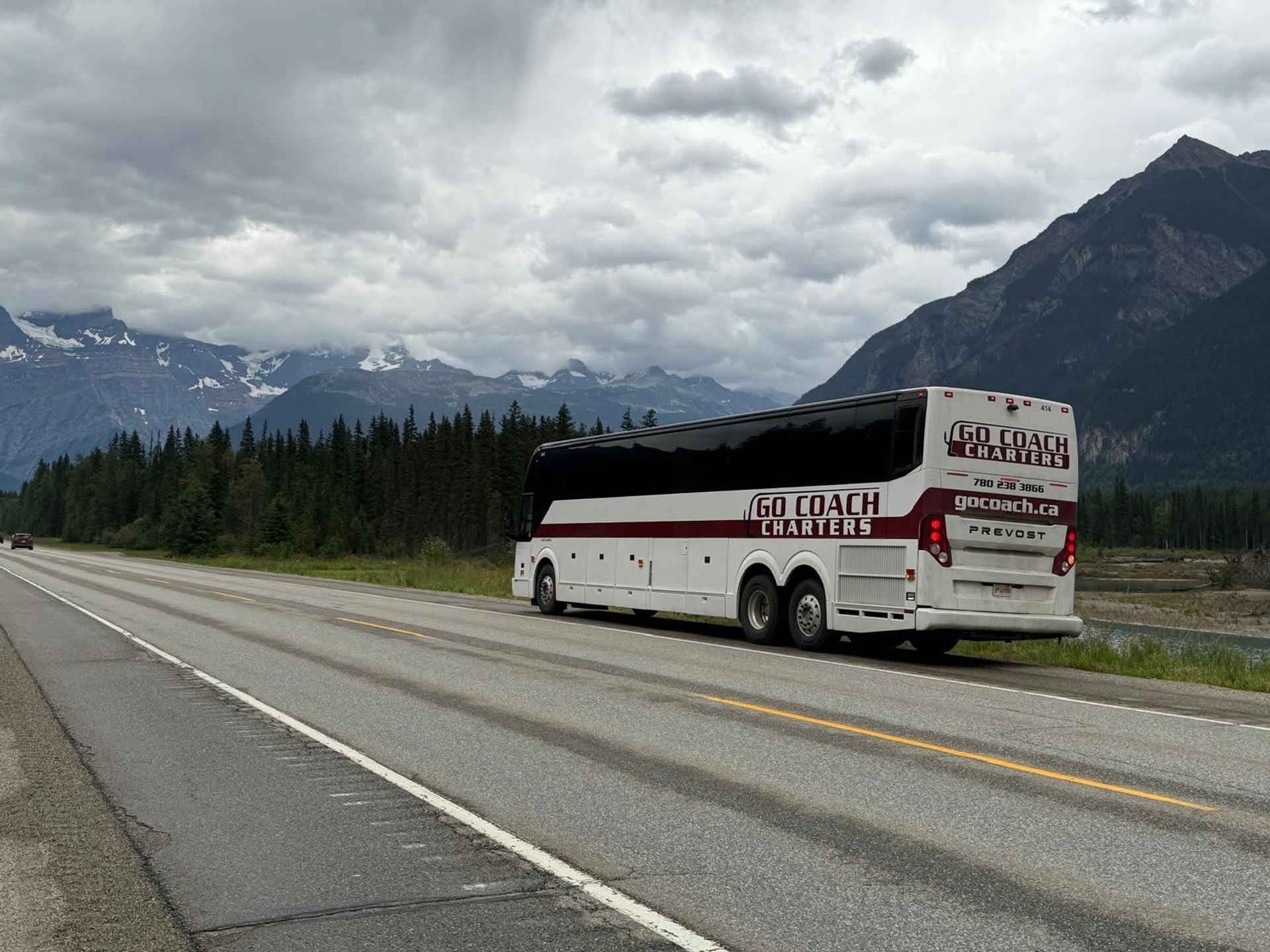 A motorcoach travels along a highway with mountains, evergreen trees, and dramatic clouds stretching across the landscape in the background.