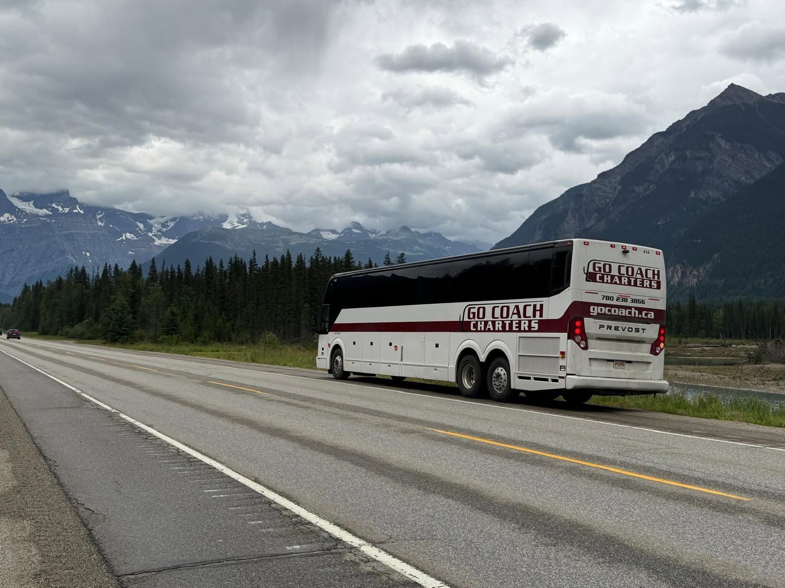 A motorcoach travels along a highway with mountains, evergreen trees, and dramatic clouds stretching across the landscape in the background.