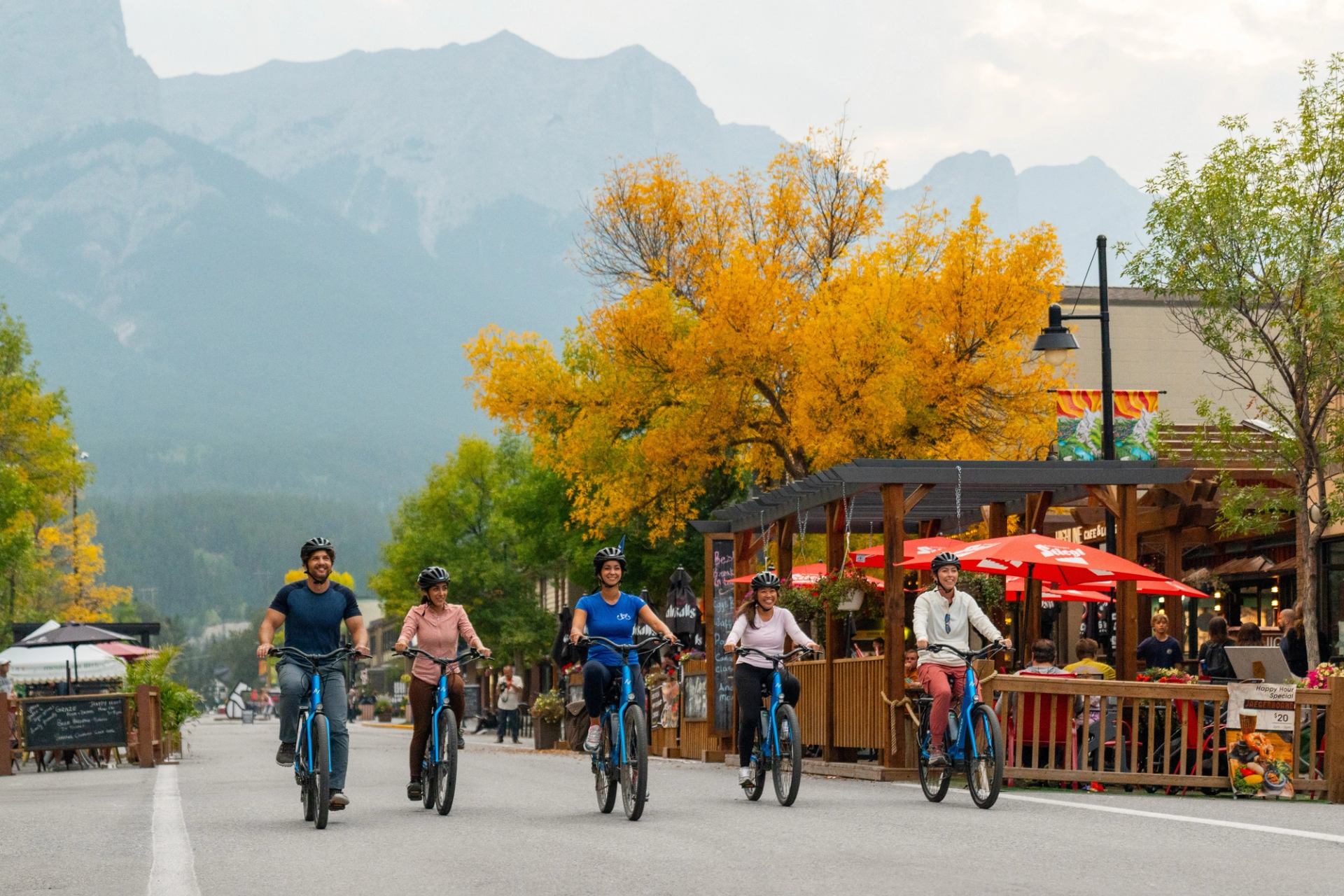Cyclists on blue bicycles ride past an outdoor patio with red umbrellas and autumn trees.