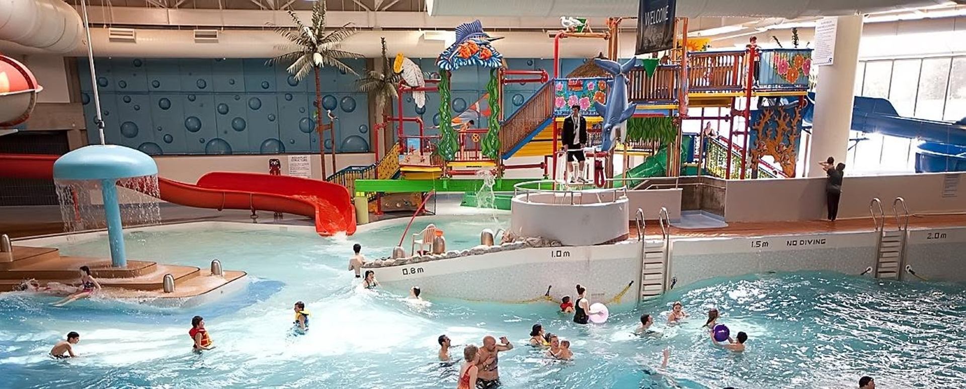 Indoor water park with wave pool, slides, splash features and people swimming.