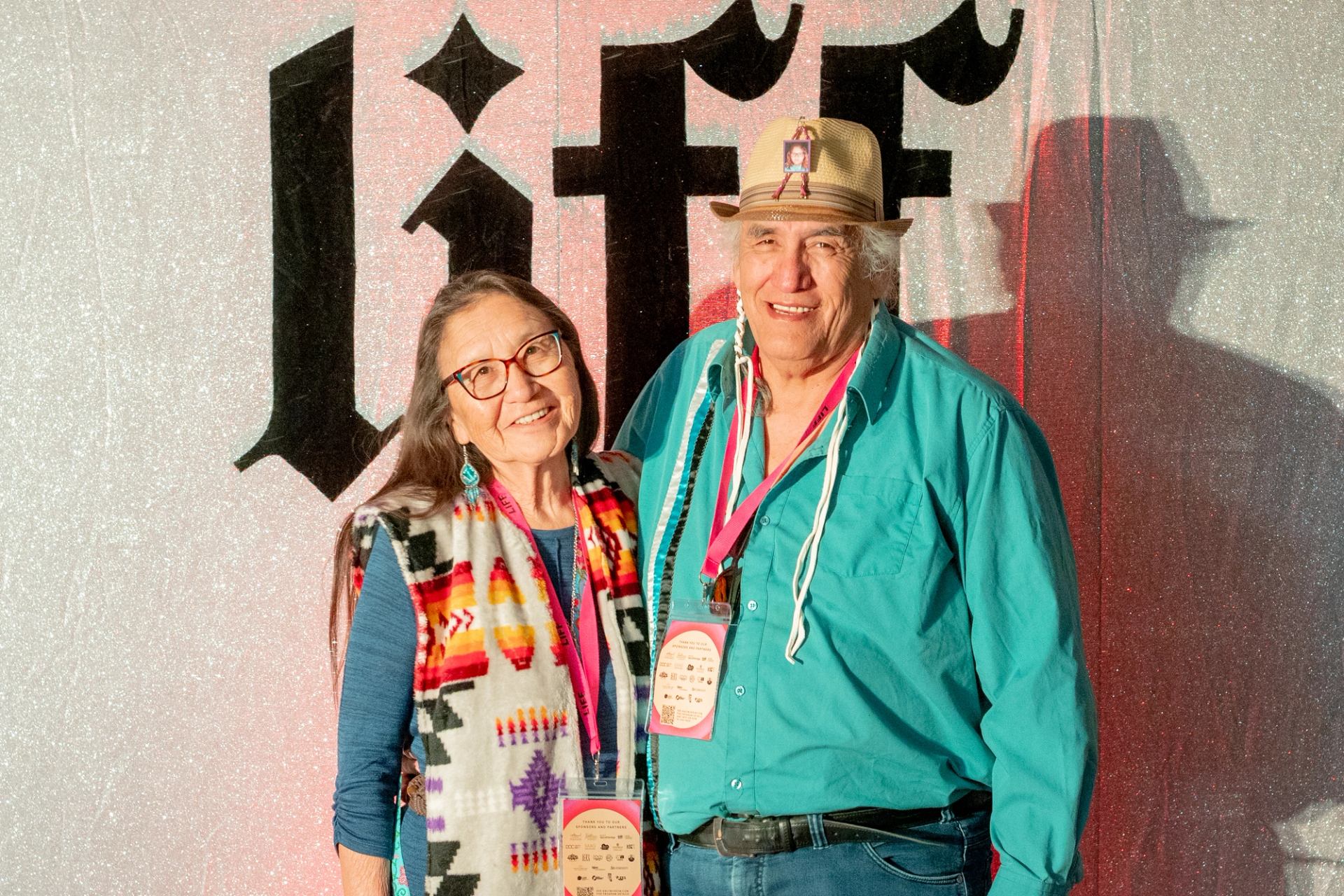 Two people with festival lanyards stand before glittery backdrop with large stylized text.