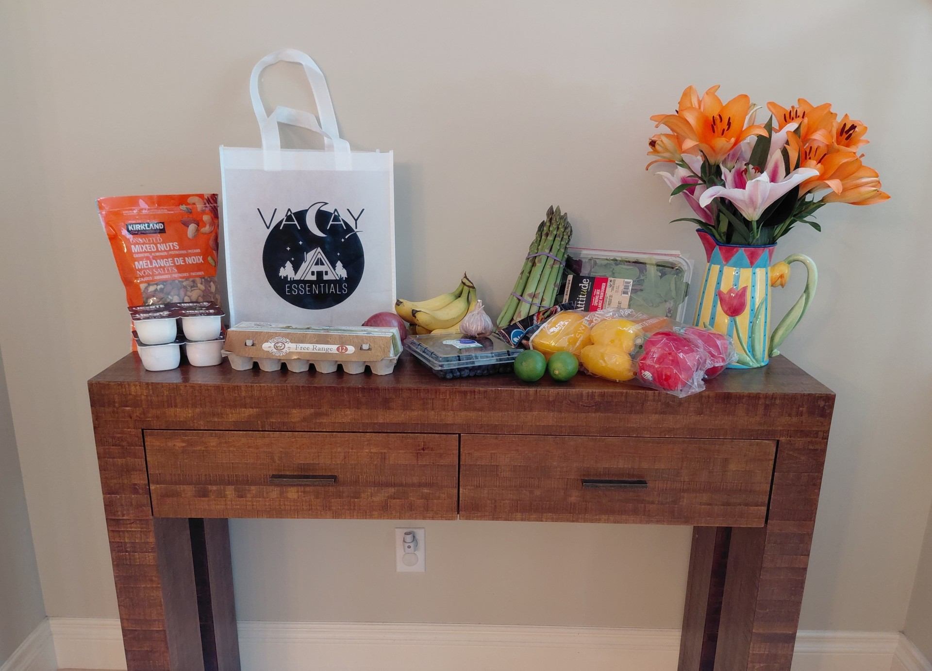 Groceries and flowers on a table with a “VACAY” bag and fresh produce.