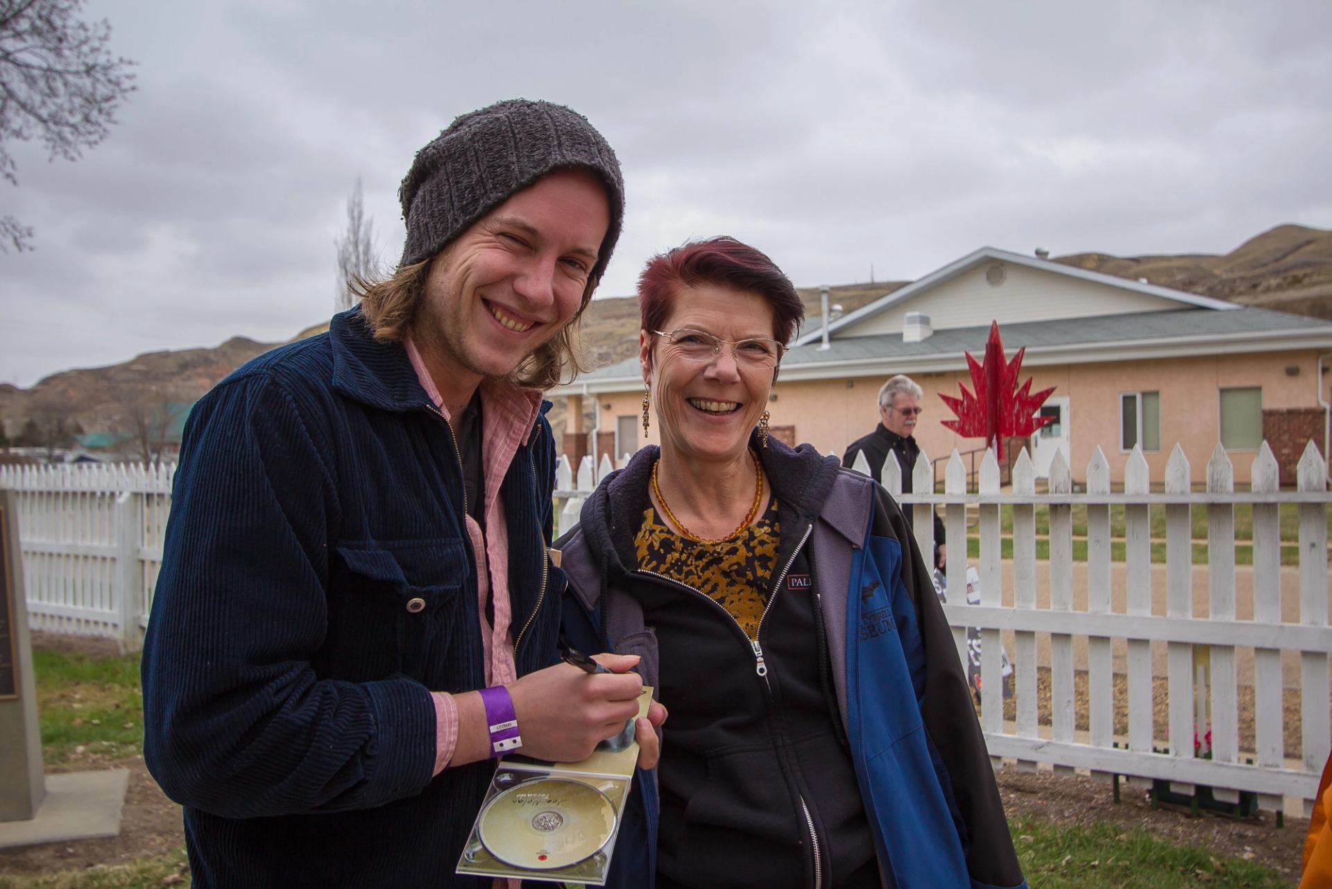 Two people standing outdoors near a white picket fence holding a CD case.