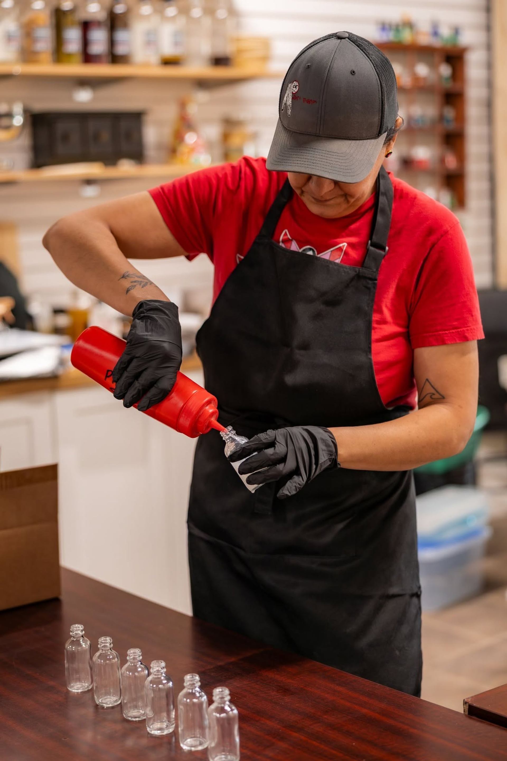 Worker in apron and gloves pours liquid into small glass bottle on table.