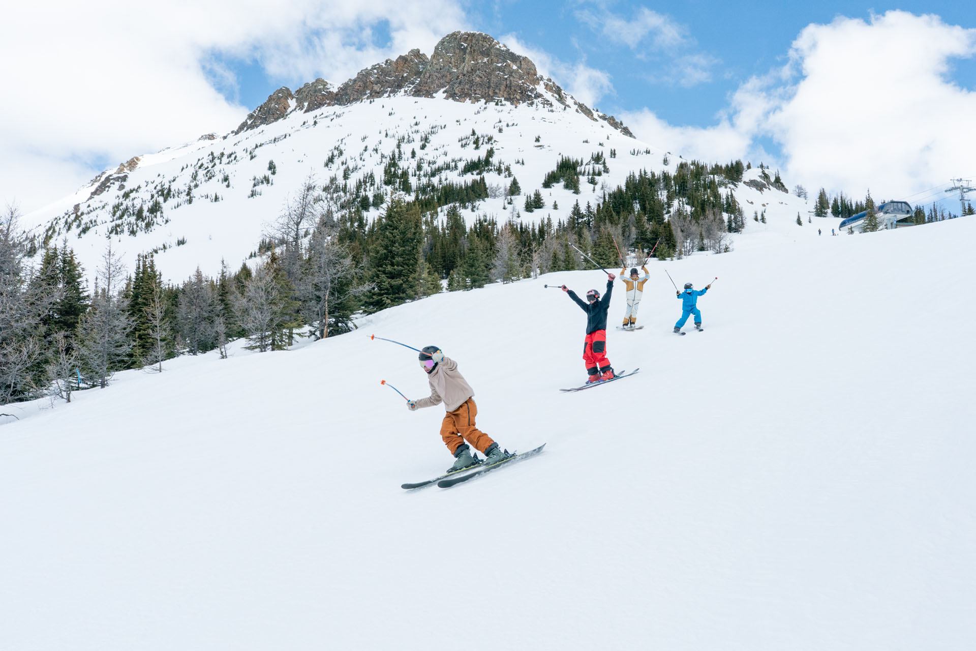Skiers glide downhill with raised poles on wide snowy slope below rocky mountain.