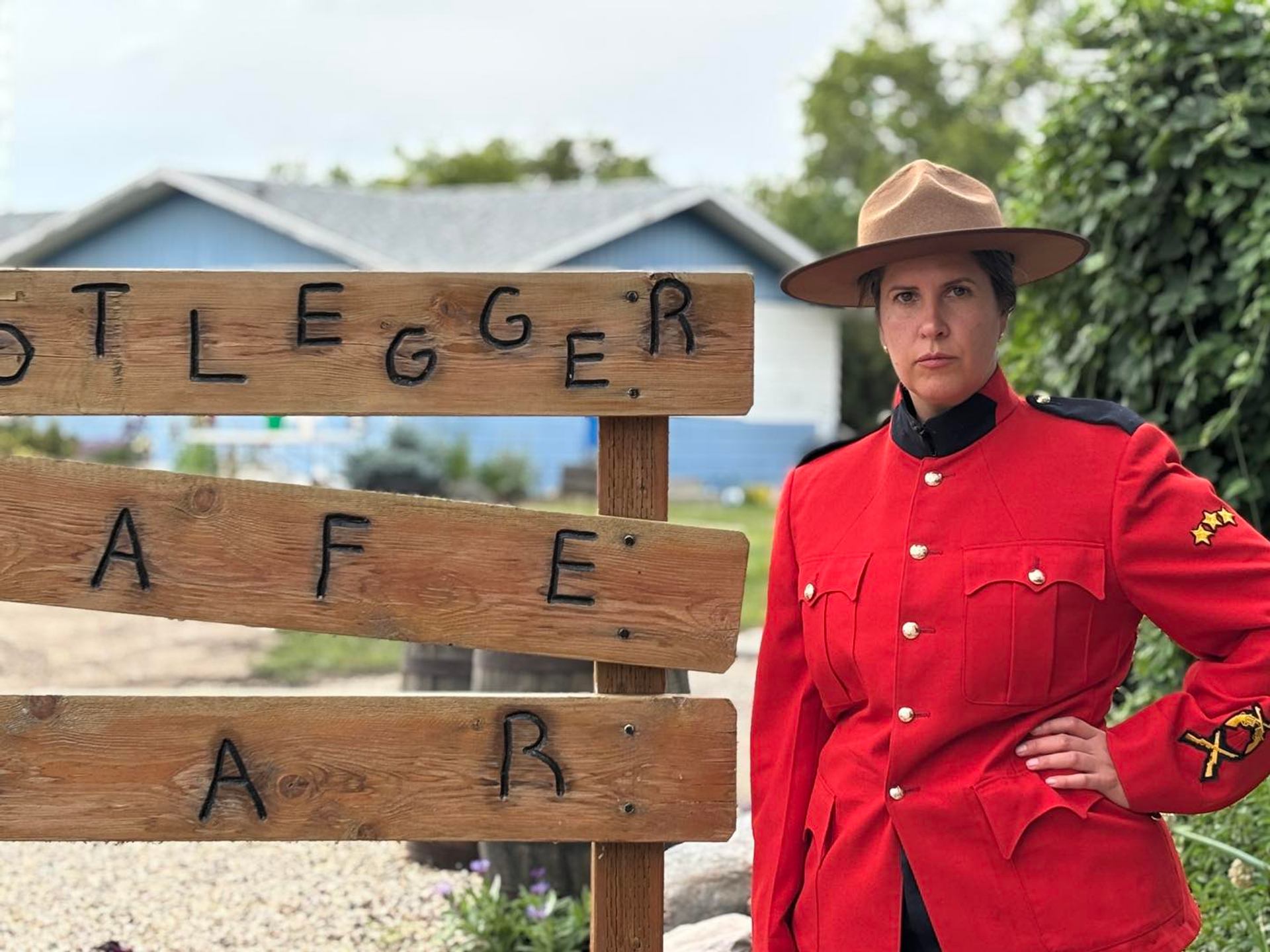 Person in red Mountie-style uniform stands beside rustic wooden sign near houses.