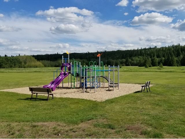 Playground with slide and climbing structures in grassy field near trees and benches.
