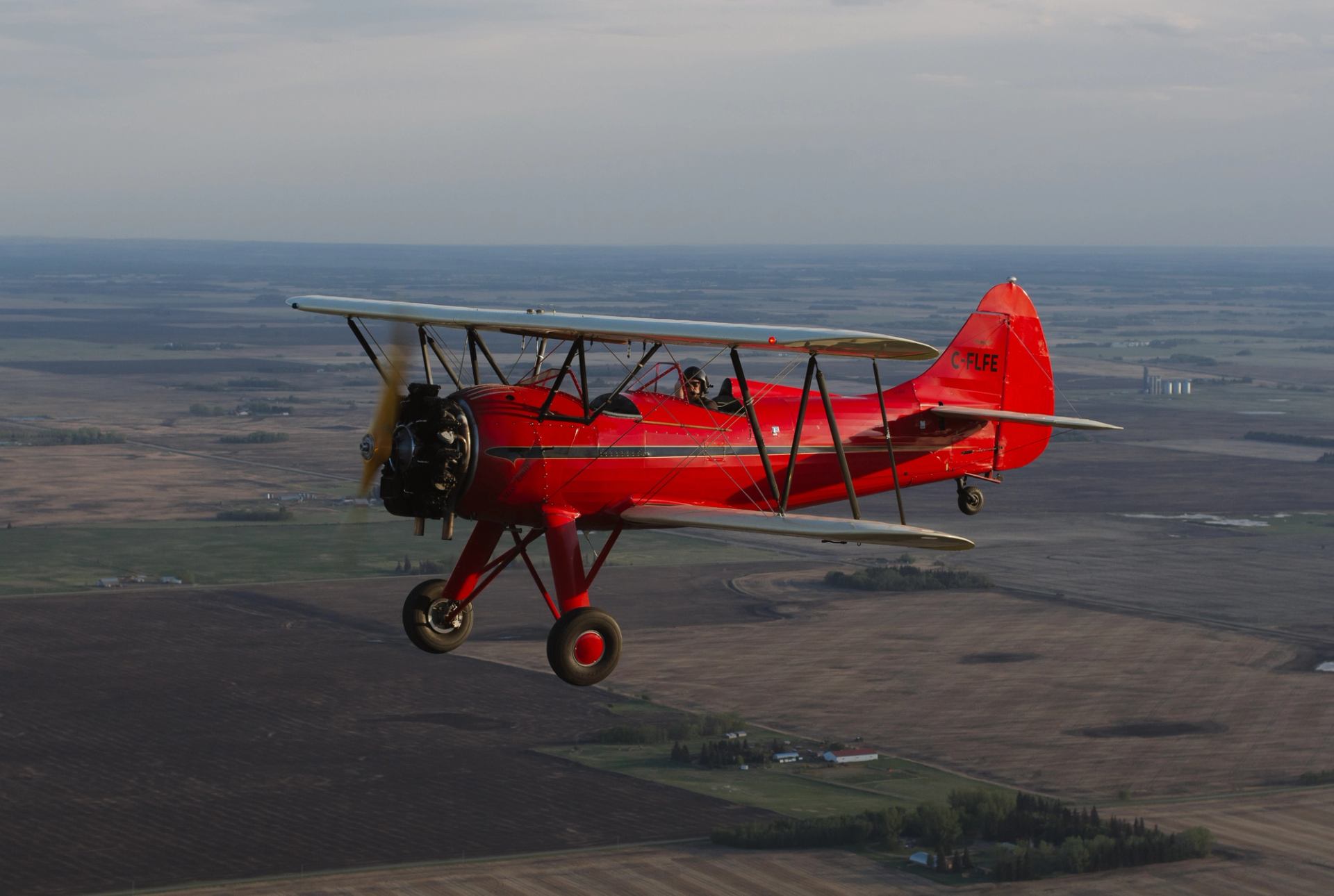 Red biplane flying above flat farmland landscape with fields and distant buildings.
