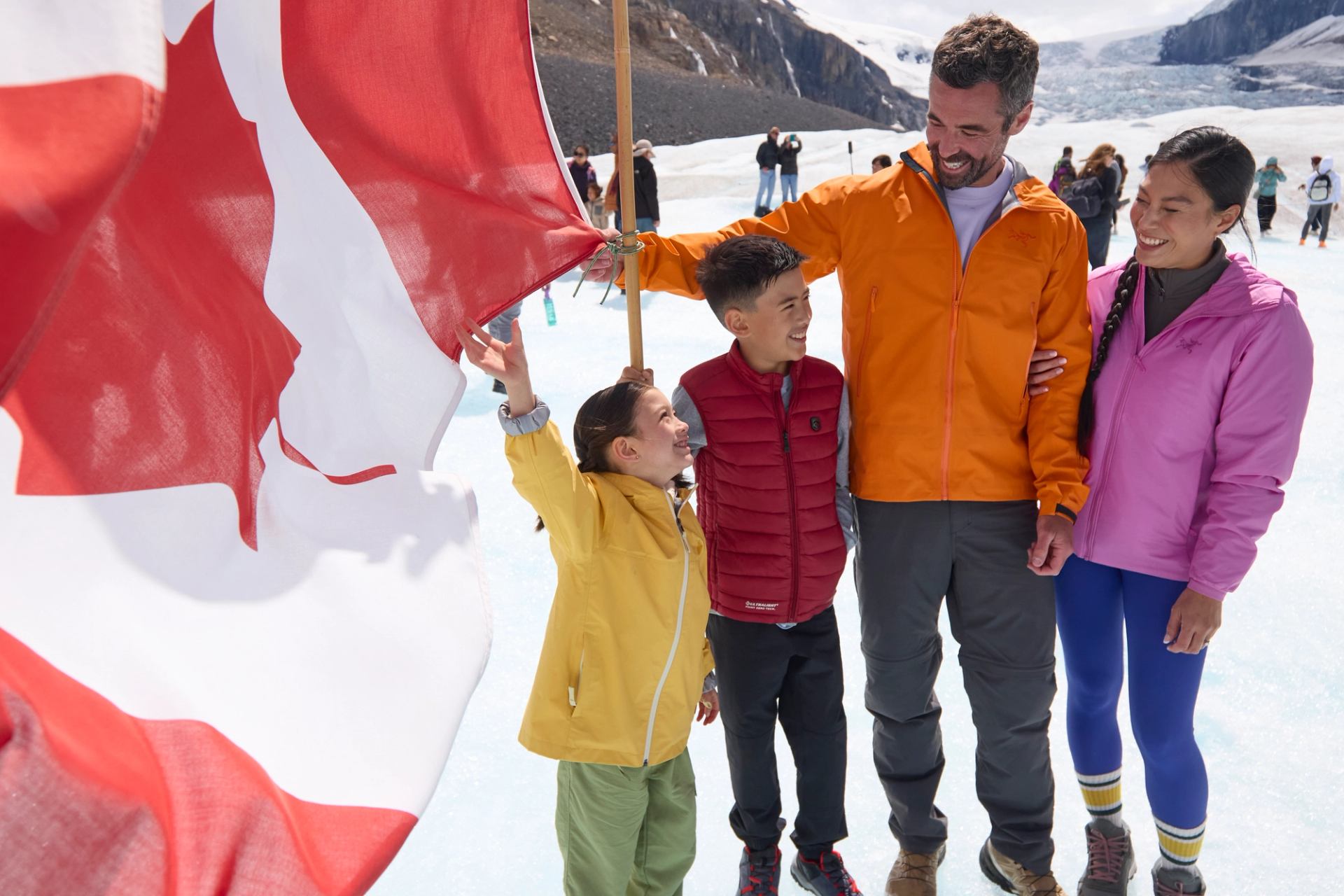 Group of people stand on snow beside large Canadian flag and distant glacier.