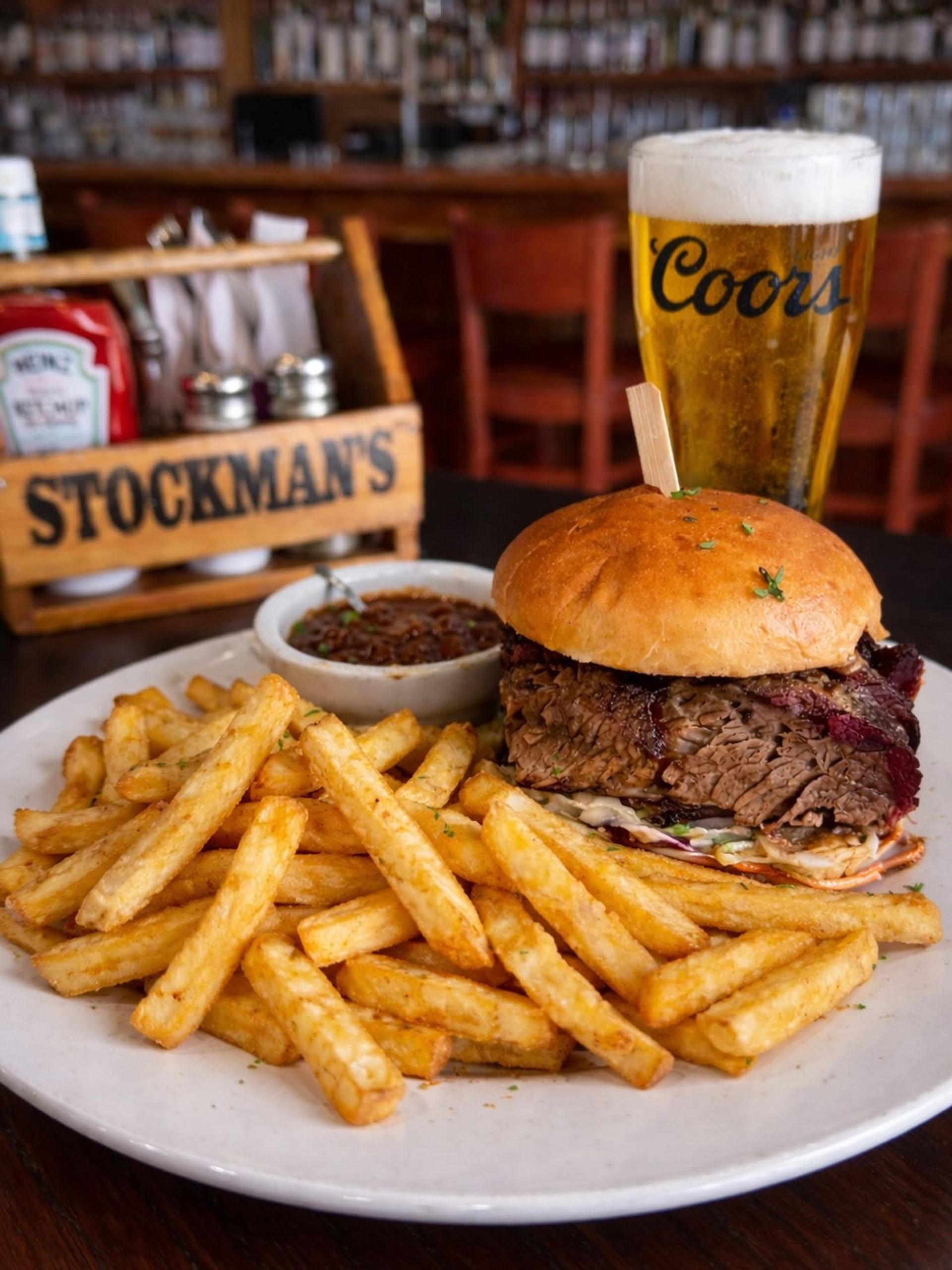 Plate of fries and brisket sandwich with dip beside Coors beer on bar table.