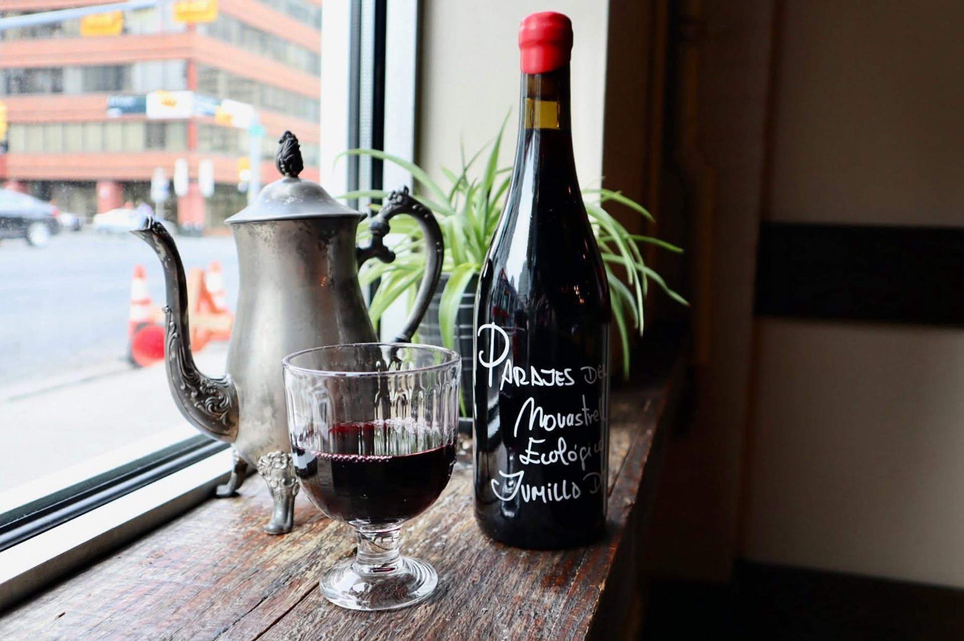A glass of red wine and bottle sit beside a vintage teapot on a sunny windowsill.