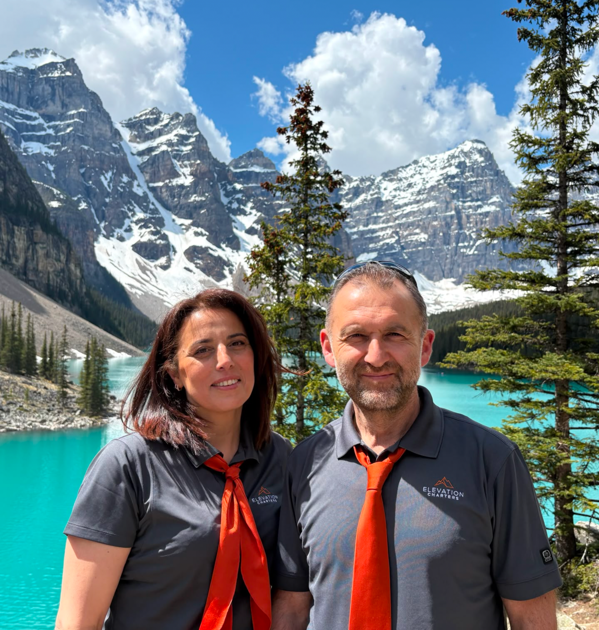 Two people in matching shirts stand by turquoise mountain lake with snowy peaks.