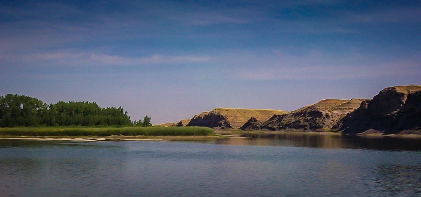 Wide river with grassy bank, trees and rocky cliffs under cloudy sky.