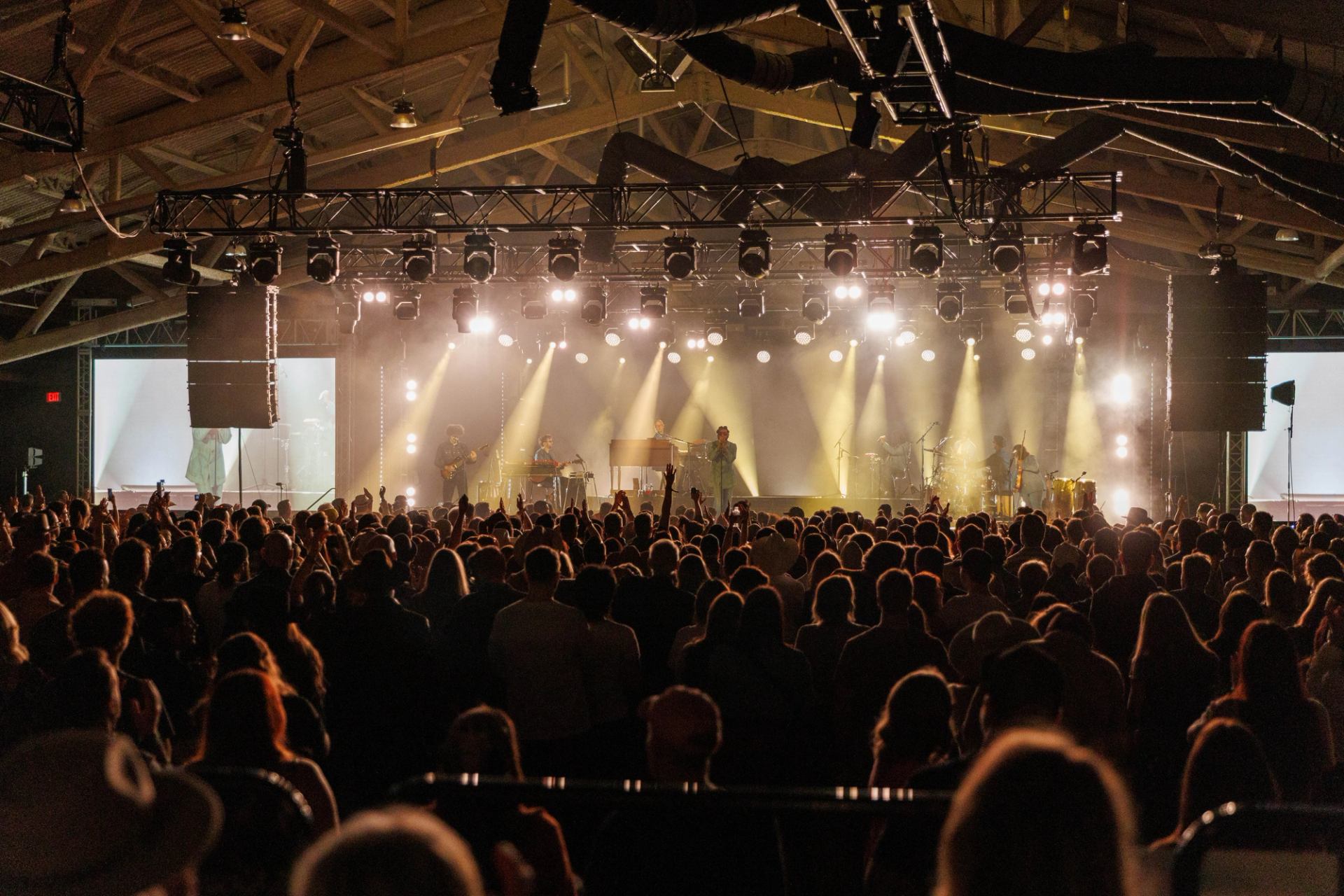Indoor concert crowd faces brightly lit stage with band performing under spotlights.