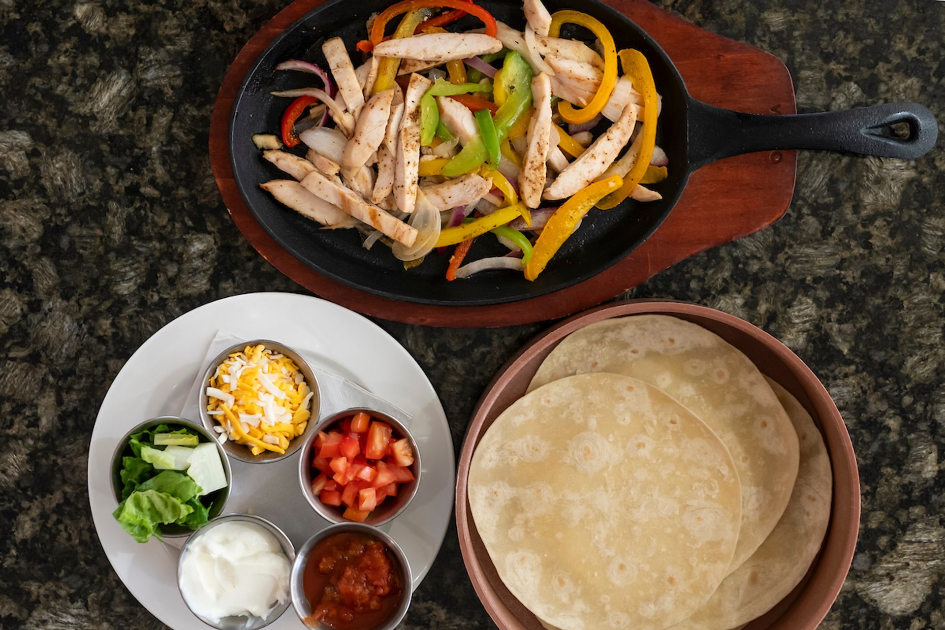 Chicken fajitas in skillet with peppers, tortillas, cheese, salsa and fresh toppings.