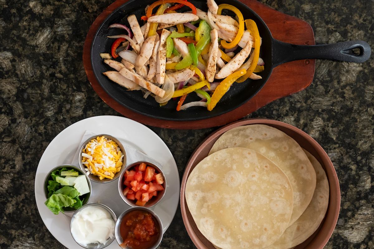 Chicken fajitas in skillet with peppers, tortillas, cheese, salsa and fresh toppings.