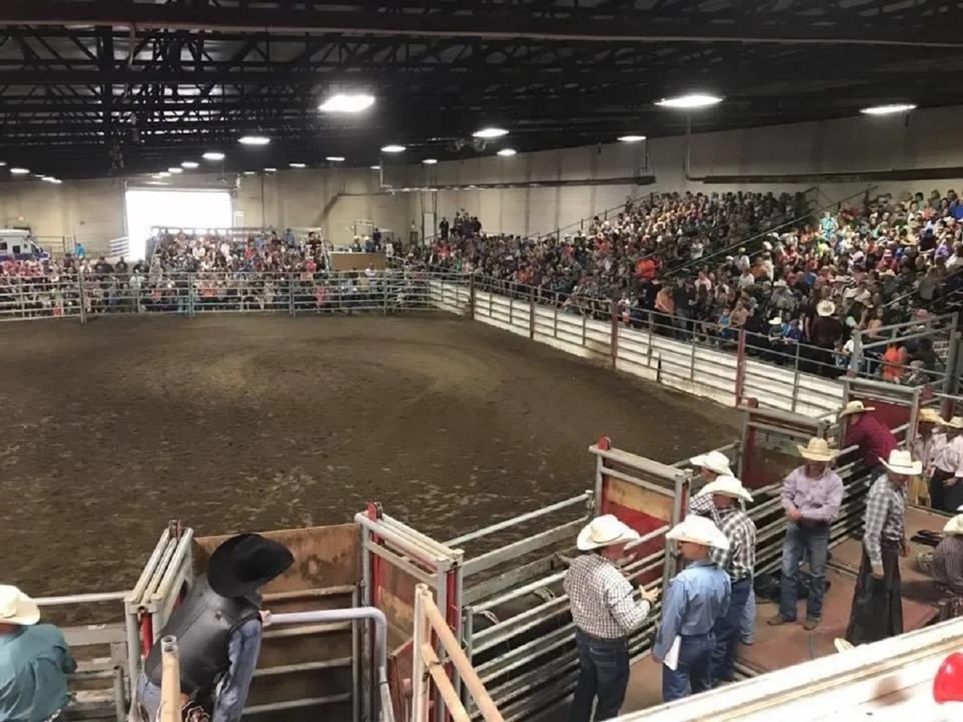 Indoor rodeo arena with a full crowd and riders preparing near the chutes.