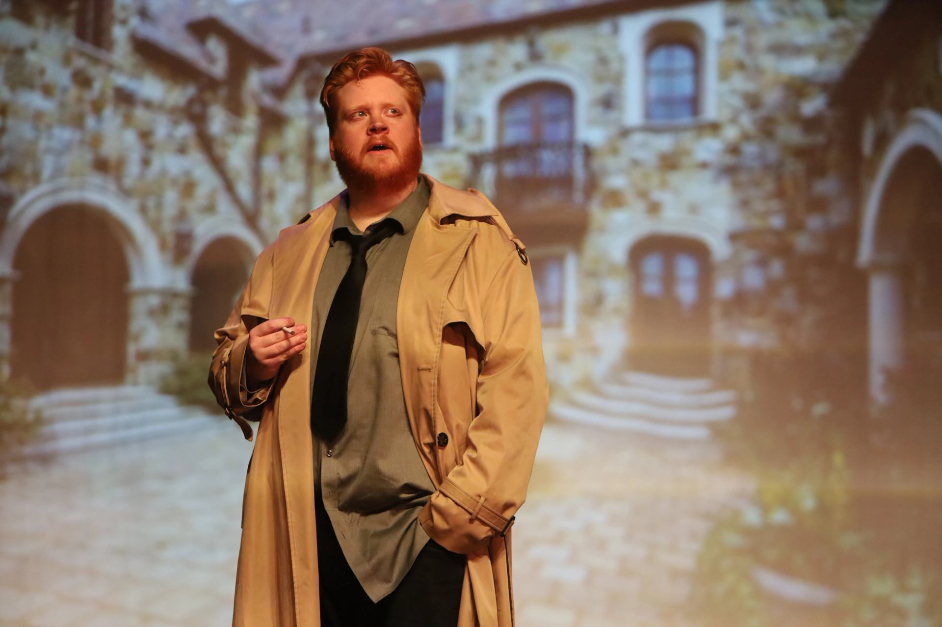 Performer in trench coat on stage with a stone building backdrop.