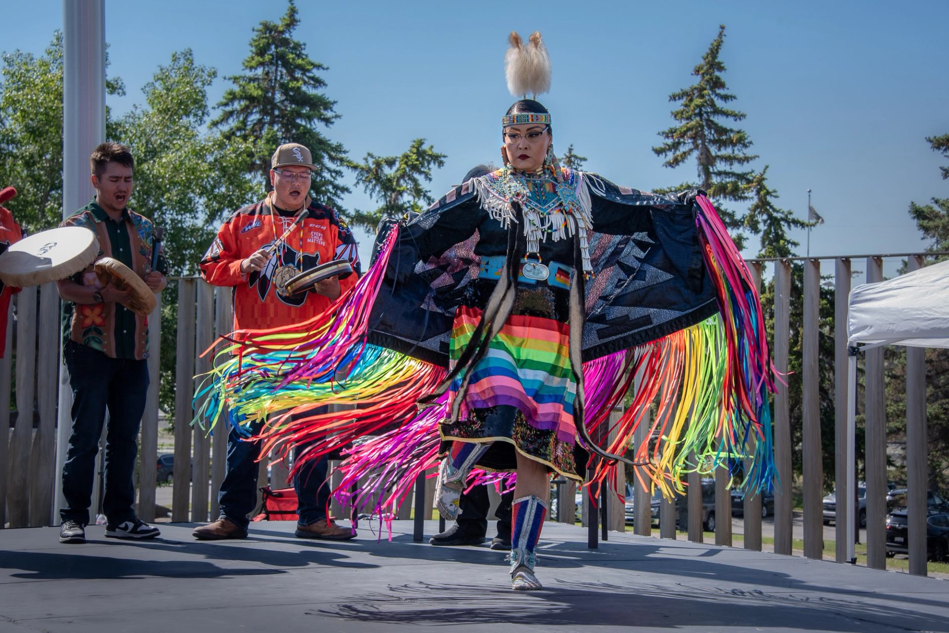 Powwow dancer in rainbow regalia performs on outdoor stage as drummers play nearby.