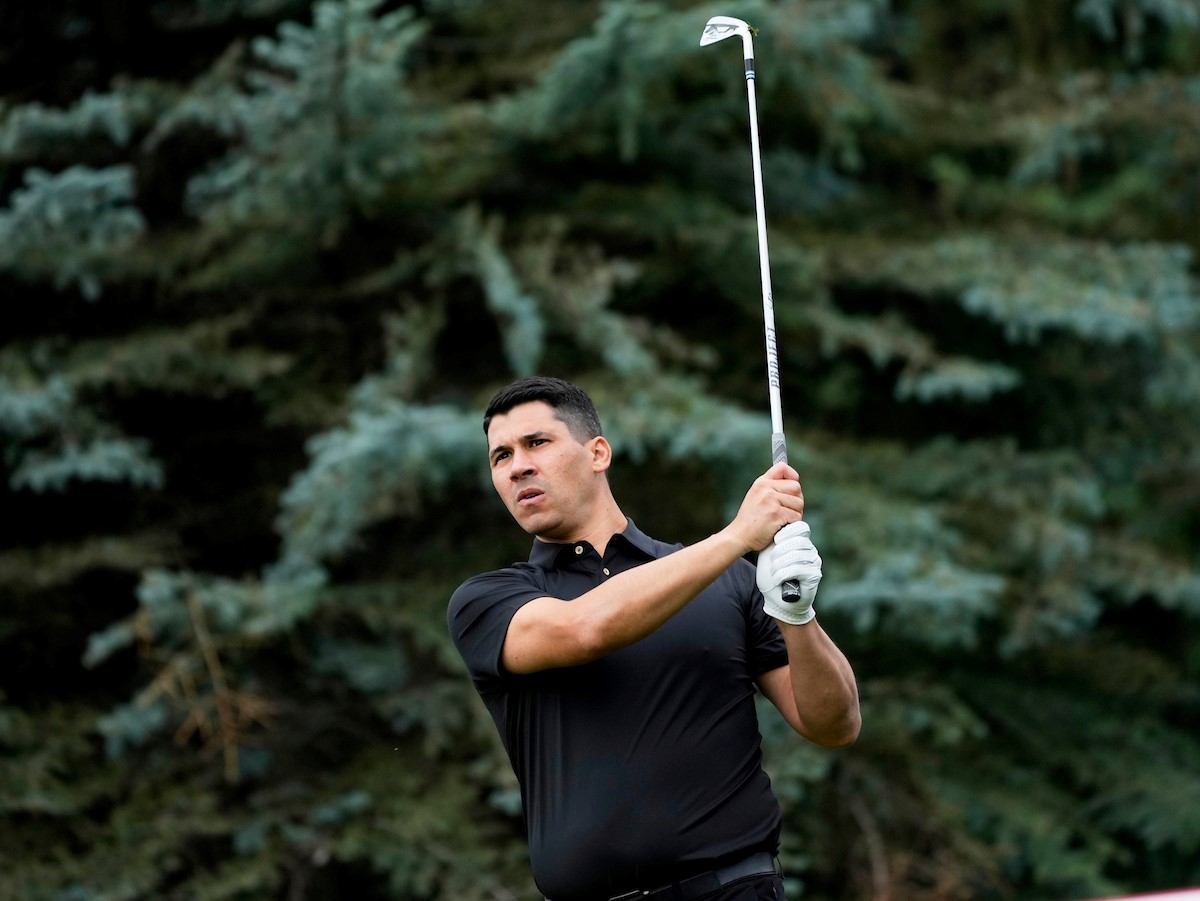 Golfer swings a club at a course, with a blurred green background behind.