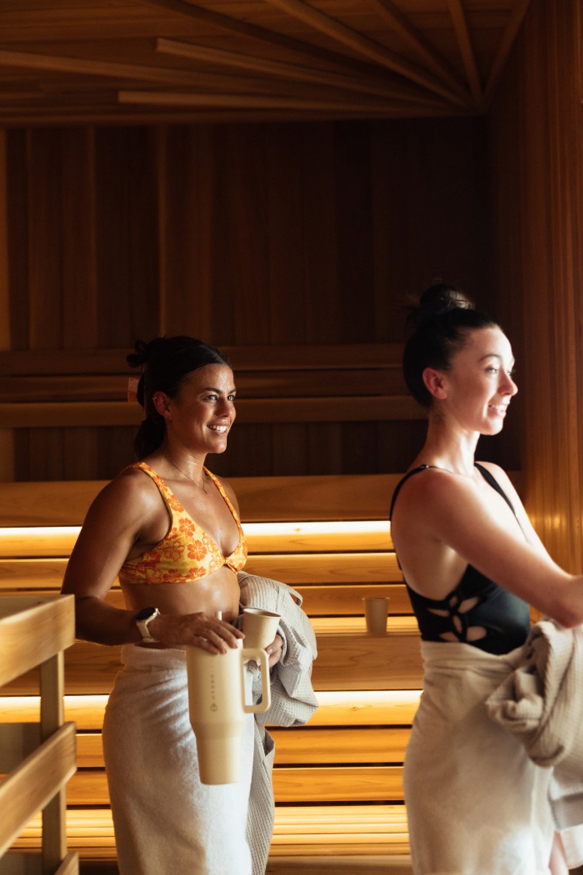 Two people in swimsuits stand in a wooden sauna, holding towels and drink containers.