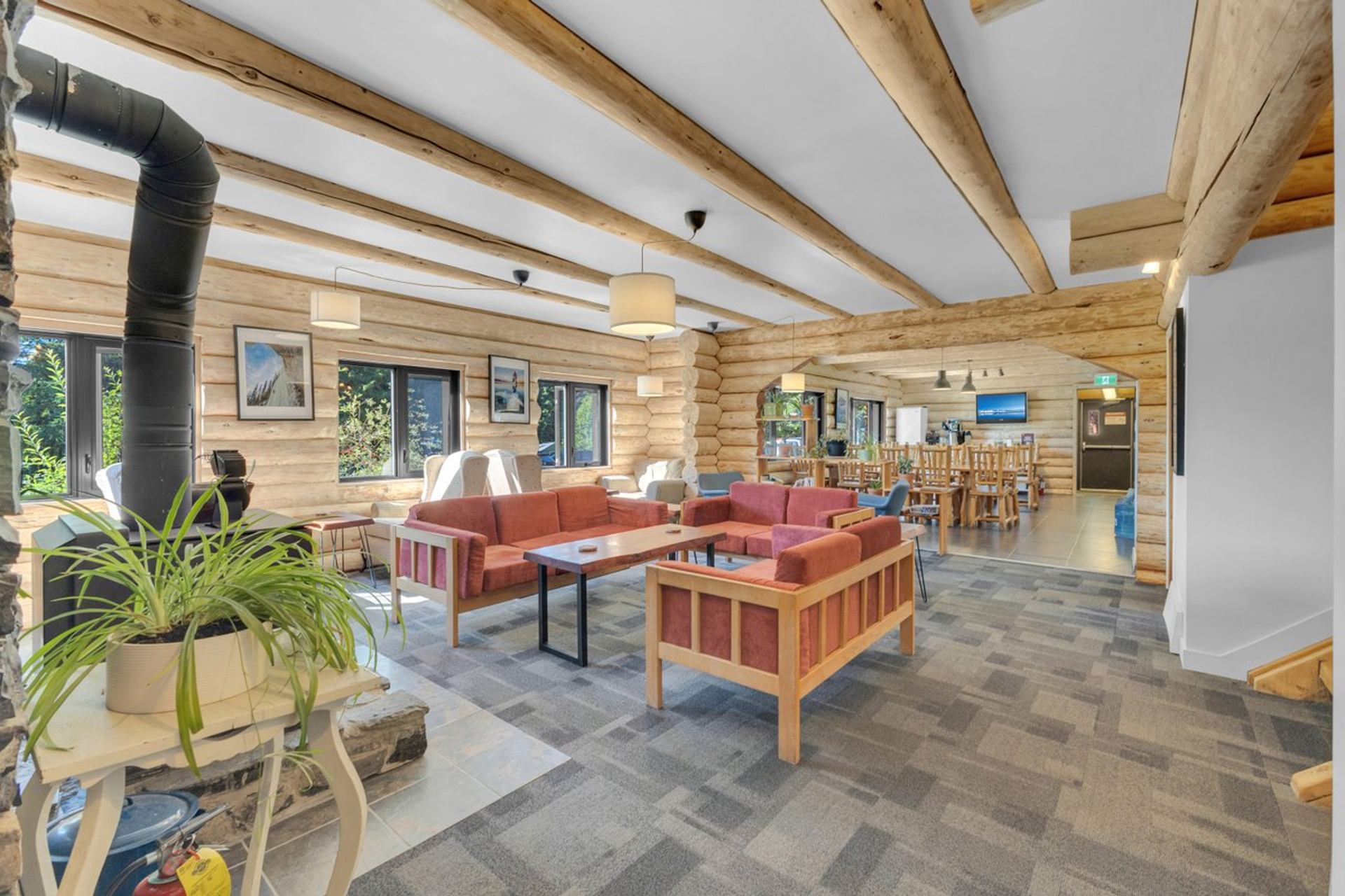 Bright lounge with wooden beams, red sofas, and a wood stove in a rustic cabin-style setting.