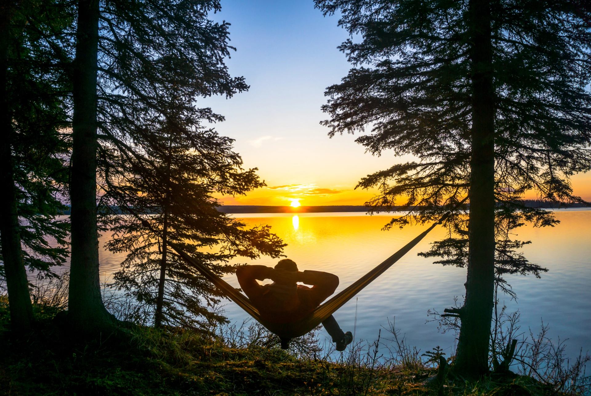 Silhouette in hammock between trees overlooking calm lake and bright sunset sky.