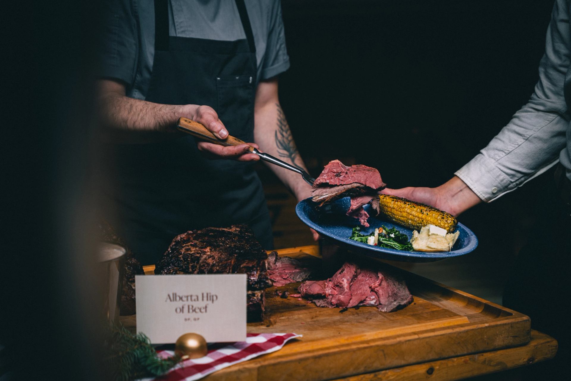 A plate being served with carved Alberta beef, corn, and greens at a rustic carving station.