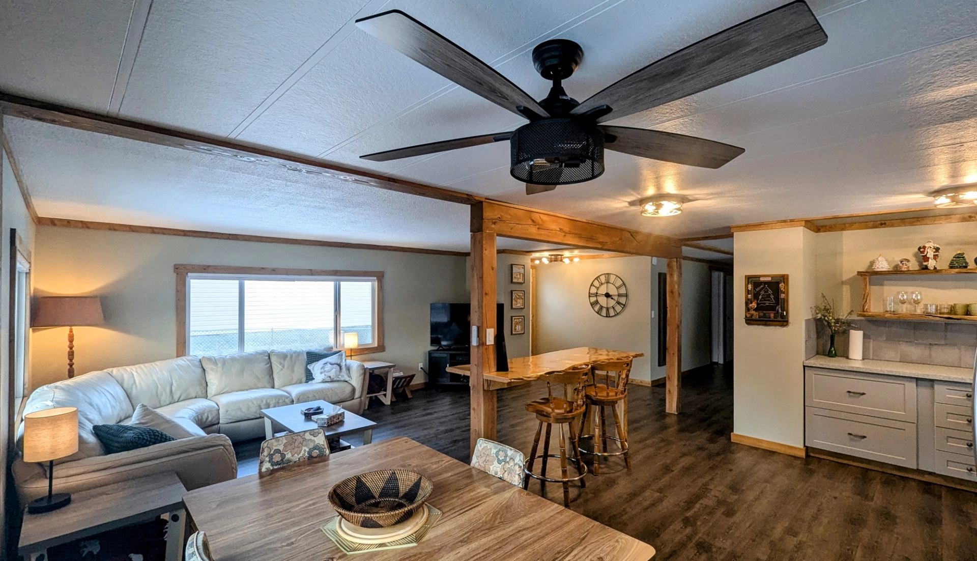 An open-concept living room, dining area, and kitchen with a sectional sofa, wooden table, and large ceiling fan.