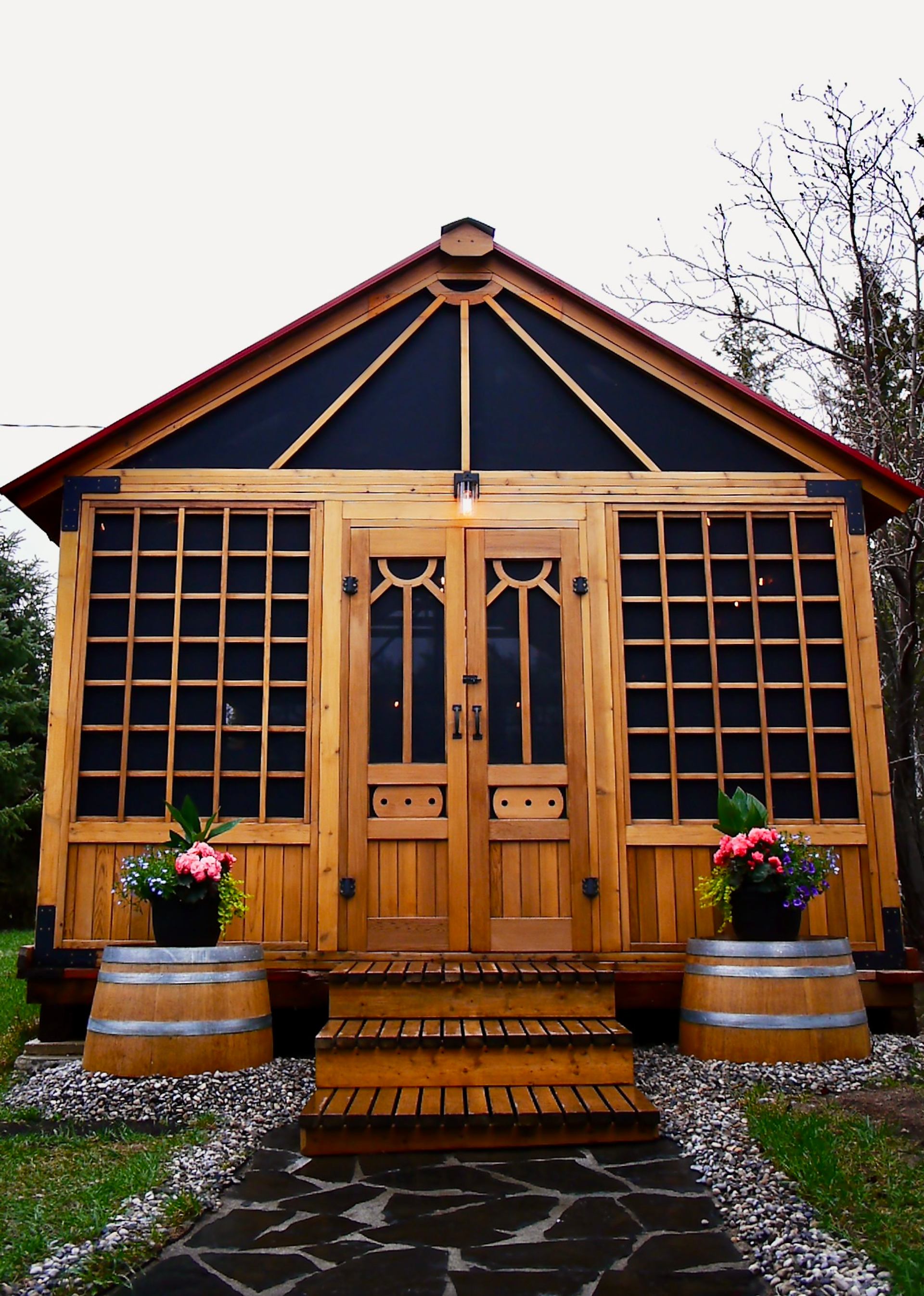 A small wood-clad sauna building with decorative paneled windows and double doors sits on a raised deck, flanked by planters. The compact structure is surrounded by trees and natural landscaping.