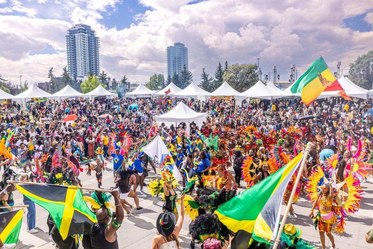 Crowd at outdoor festival with colourful costumes, Caribbean flags and white vendor tents.