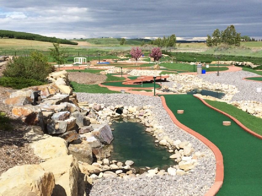 Mini-golf course with rock-lined stream, footbridge and winding greens under cloudy sky.