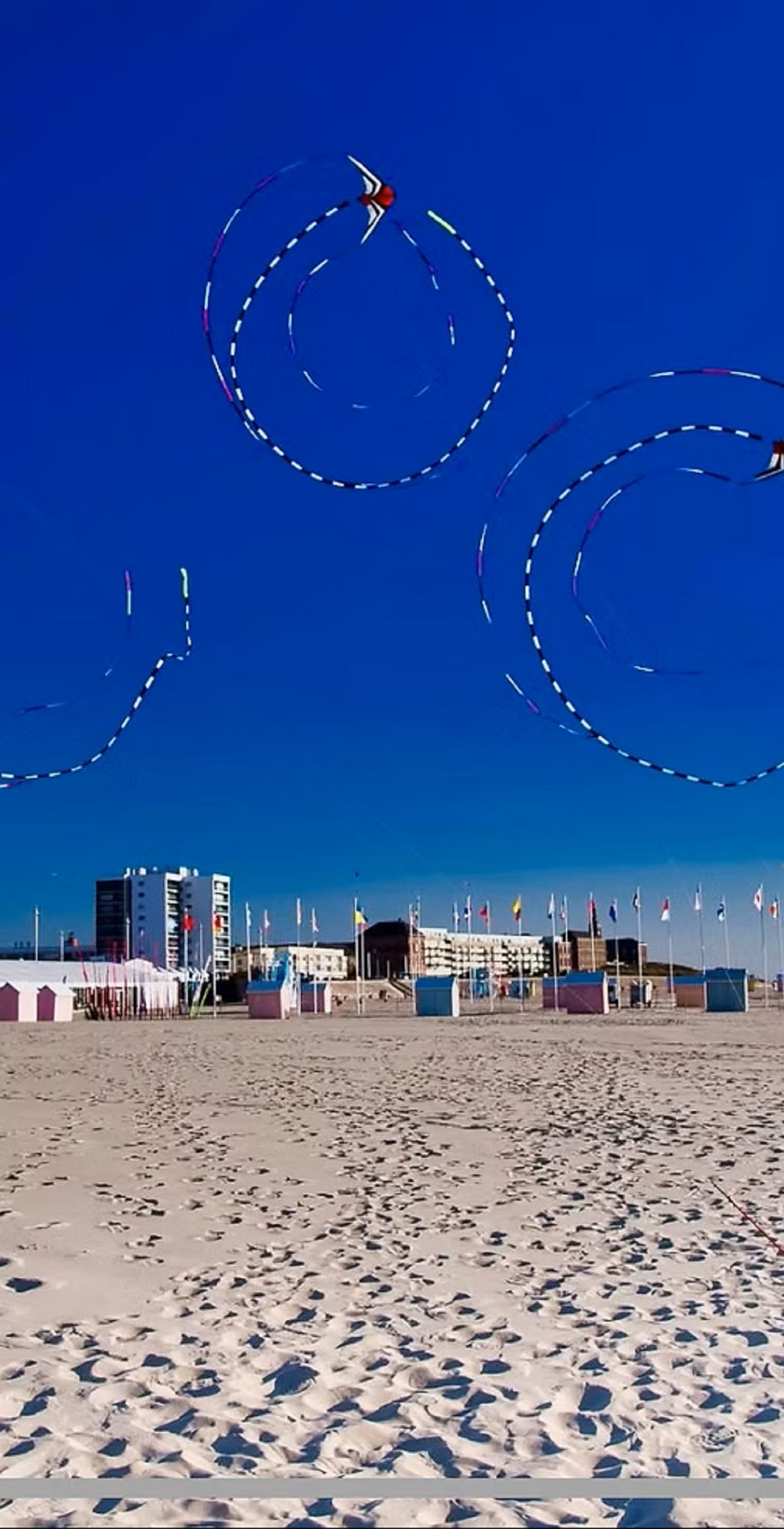 Colourful kites loop above sandy beach with beachfront buildings and flagpoles in background.