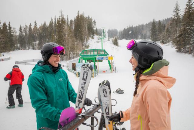 Two skiers retrieve their skis from the rack at the base of the hill, chairlift in the background, before skiing Hidden Valley Ski Resort.