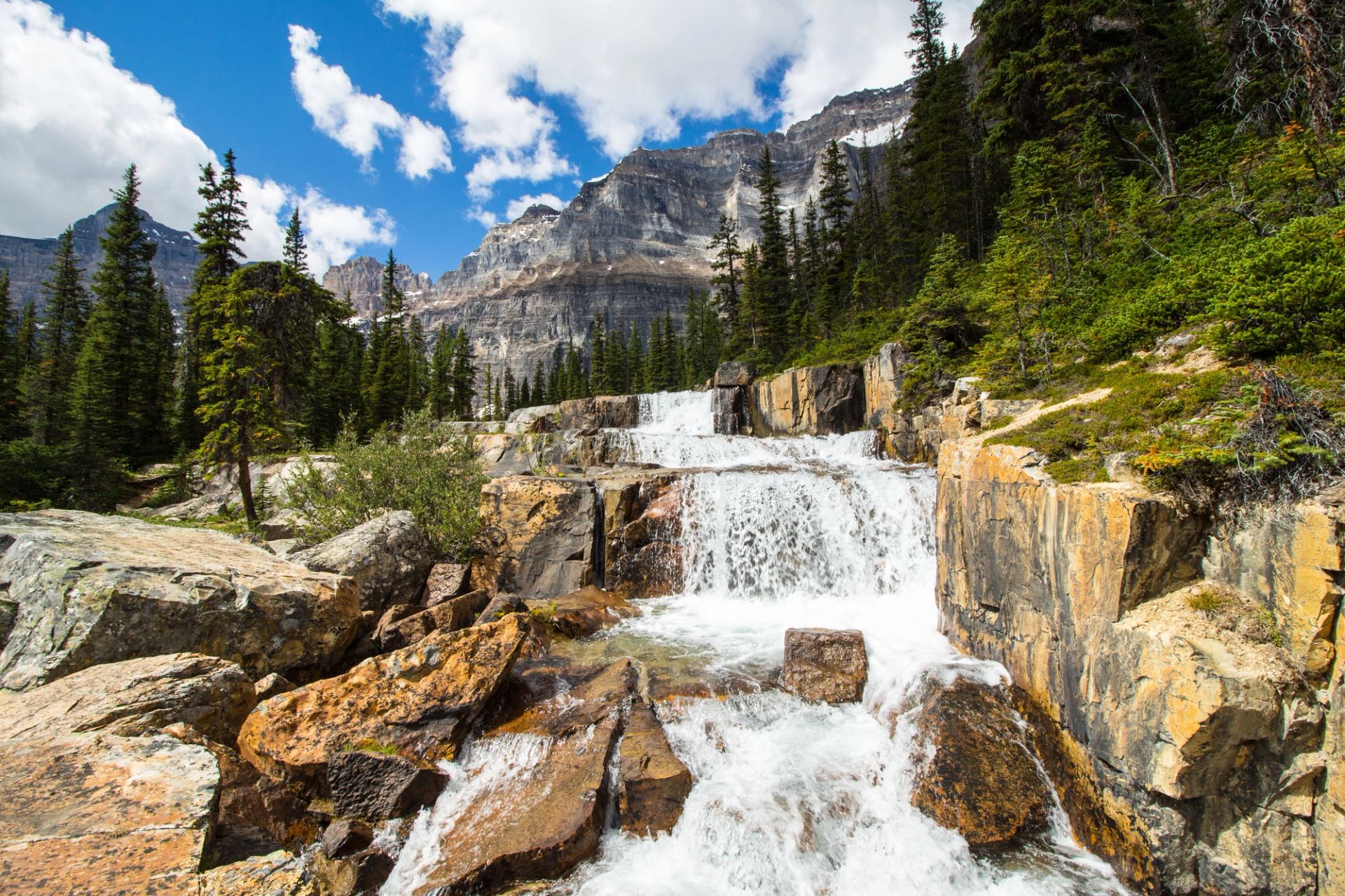 Mountain waterfall cascading over rocky ledges, surrounded by evergreen trees and distant cliffs.