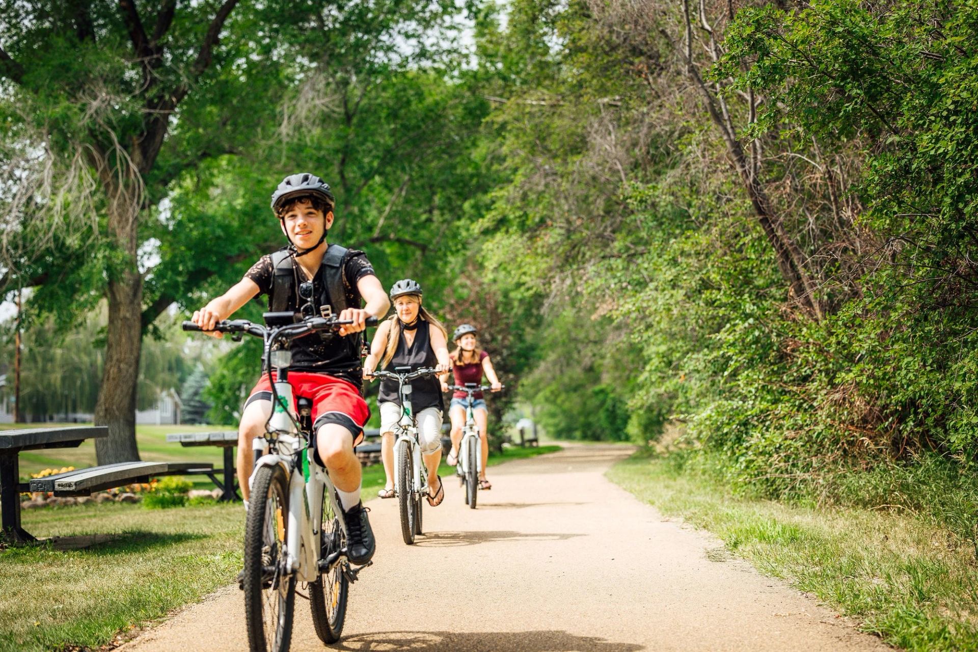 Three young people ride bikes on a tree-lined paved trail in a sunny park with picnic benches.