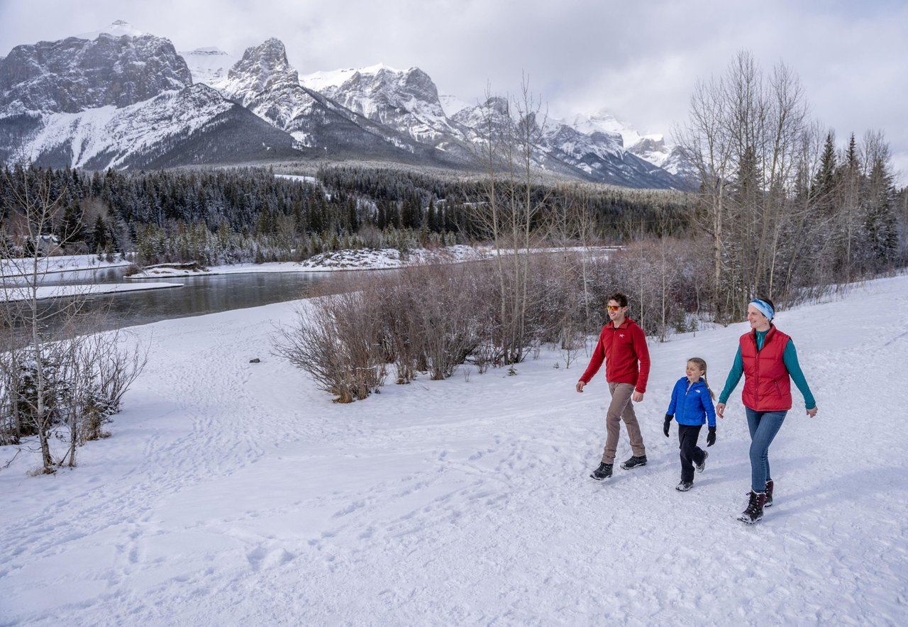 A family (couple and young child) walking the Bow River Loop Trail in the winter, on the snow-covered path, with mountains in the background