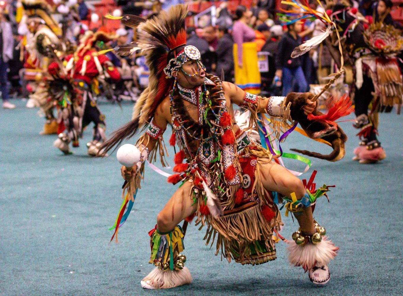 Powwow dancer in colourful regalia performs on arena floor before seated crowd.
