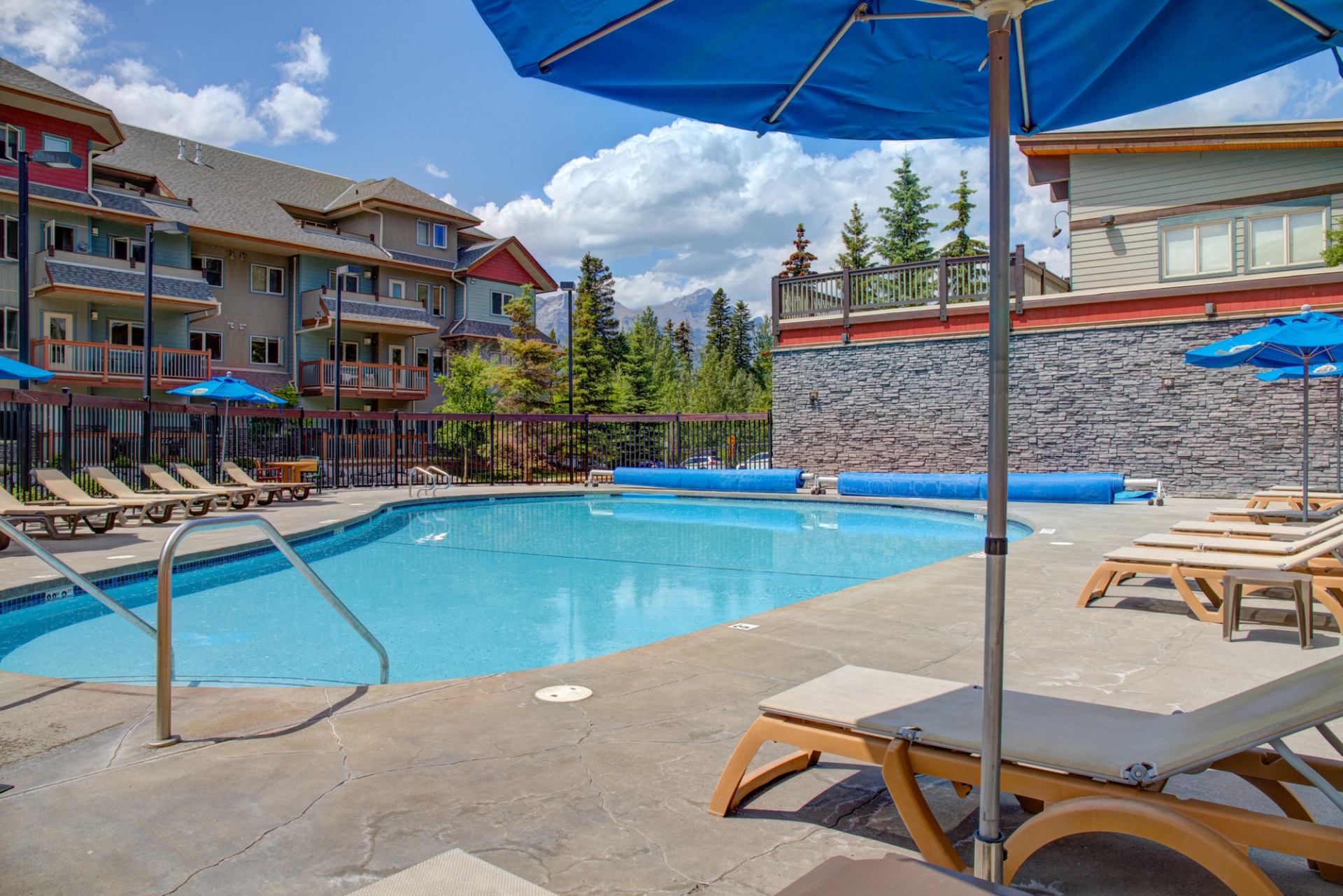 Outdoor pool area with lounge chairs, blue umbrellas, and mountain lodge backdrop.