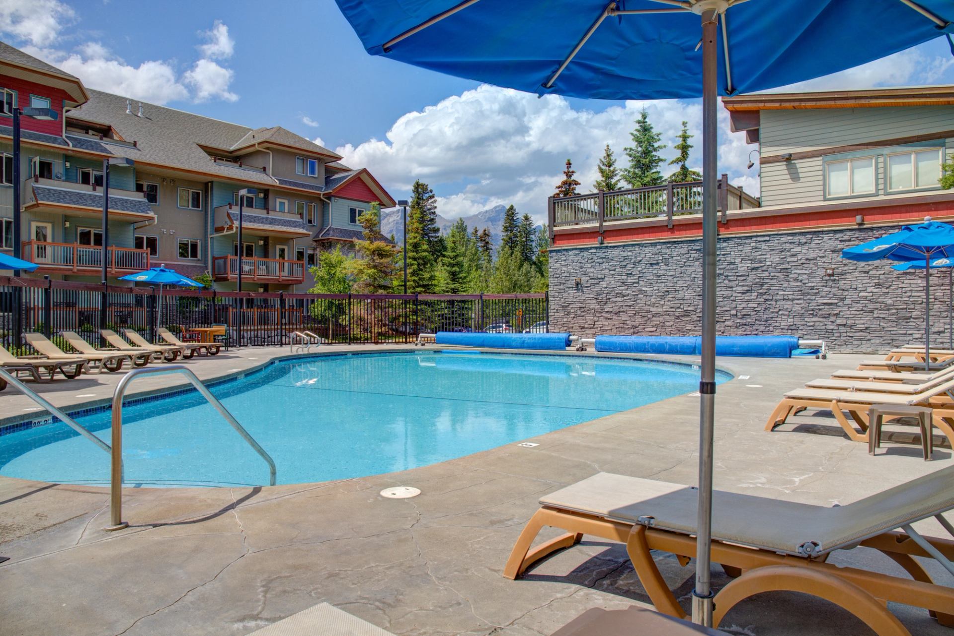 Outdoor pool area with lounge chairs, blue umbrellas, and mountain lodge backdrop.