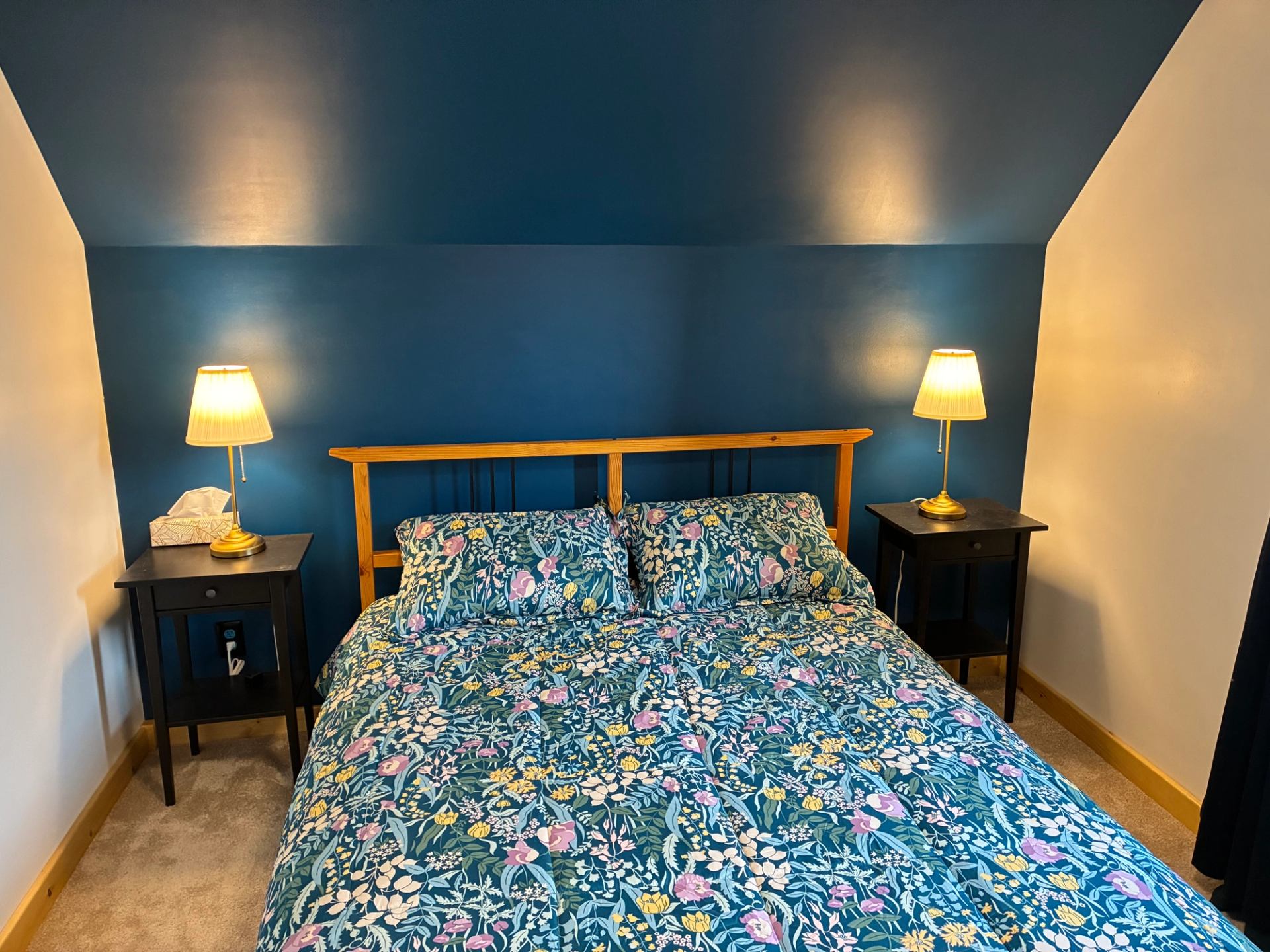 Bedroom with floral bedspread, wooden headboard, blue accent wall and two lit lamps.