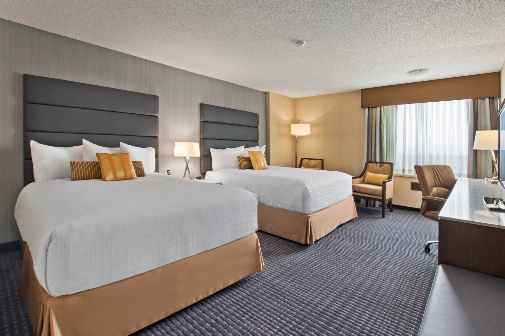 Hotel room with two double beds, armchairs, desk, lamps and large curtained window.