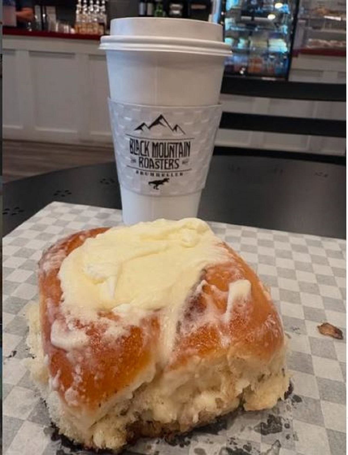 Hot coffee in a branded cup with a glazed pastry on a checkered paper inside a café.