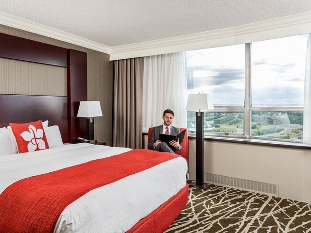 Modern hotel room with red accents, large windows, and a seated guest.