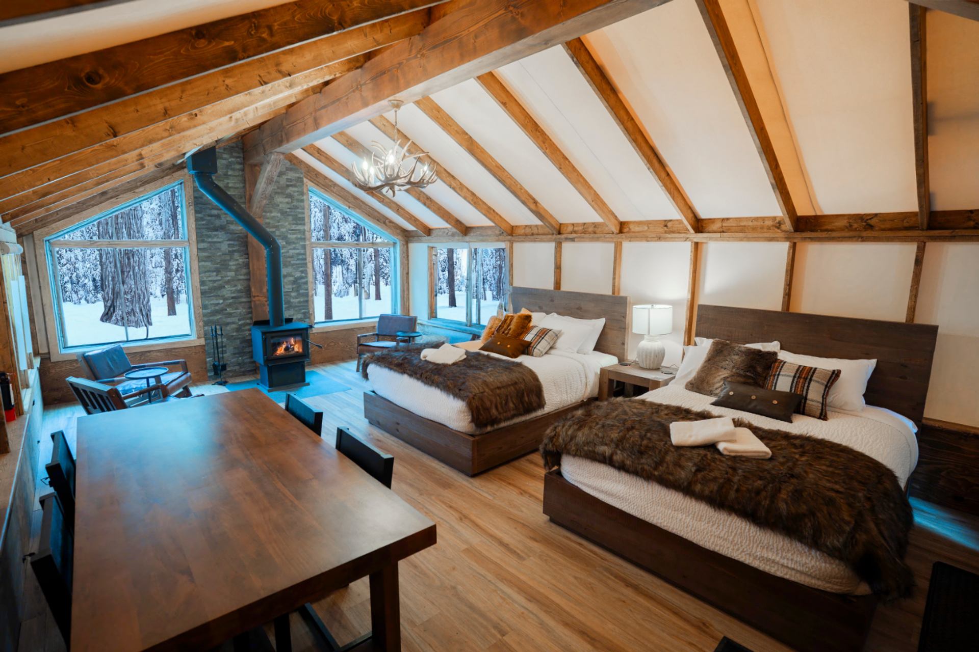 Lodge-style bedroom with two beds, wood stove, dining table and snowy forest view.